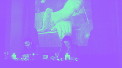 deux hommes lisent dans une salle sombre avec des bougies et une peinture projetée derrière